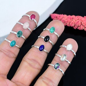 May include: A collection of silver rings, each featuring an oval gemstone in various colors, including red, pink, green, and blue. The rings are displayed on fingers, showcasing the different stone options available.