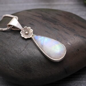 May include: A silver pendant necklace featuring a pear-shaped moonstone. The moonstone has a pearlescent, iridescent quality. The pendant is accented with a small flower and hangs from a delicate silver chain. The necklace is displayed on a dark, textured surface.