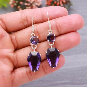 Può includere: Un paio di orecchini in argento con un design pendente. Ogni orecchino presenta una piccola gemma rotonda e viola nella parte superiore, collegata a una gemma più grande, a forma di bara, viola scuro sottostante.