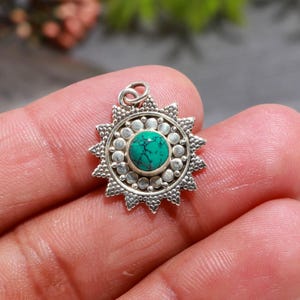 Könnte beinhalten: Ein silberner Anhänger mit Sonnenstrahlen-Design. Der Anhänger hat einen zentralen, runden, türkisfarbenen Stein, der von einem detaillierten, strukturierten Rand umgeben ist. Der Anhänger hat oben eine kleine Schlaufe für eine Kette.