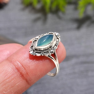 Puede incluir: Anillo de plata con una piedra preciosa azul claro en forma de marquesa. La piedra está engastada en un bisel ornamentado con un diseño floral. La banda del anillo es delgada y dividida en los hombros.