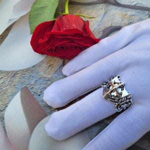 May include: A silver-toned ring with an ornate design featuring a castle-like structure and small embellishments. A vibrant red rose rests nearby. The ring is displayed on a white surface.