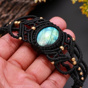 May include: Black macrame bracelet featuring a central oval labradorite stone with blue and green iridescence. The bracelet is adorned with small gold-colored beads, creating a bohemian and handcrafted aesthetic.