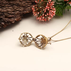 May include: Openwork gold-colored locket pendant necklace. The locket is round with an intricate floral design. The necklace chain is thin and gold-colored. The locket is open, revealing the interior. The background includes a piece of wood and pink flowers.