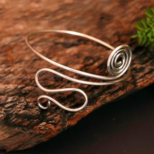May include: A silver-colored arm cuff with a spiral design and a curved, flowing shape. The cuff is made of a thin, shiny metal and rests on a dark, textured surface. The design features a spiral at one end and a decorative curve.