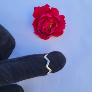 Può includere: Un anello a zig-zag d'argento è mostrato su uno sfondo nero, con una rosa rosso vivo sullo sfondo. Il design dell'anello presenta una serie di angoli acuti, creando un'estetica moderna e geometrica. La rosa aggiunge un tocco di bellezza naturale.