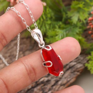 Puede incluir: Un collar de plata con un colgante de piedra preciosa roja rectangular y facetada. El colgante mide aproximadamente 2,5 cm de largo y cuelga de una delicada cadena de plata. La piedra preciosa tiene un aspecto vibrante y translúcido.