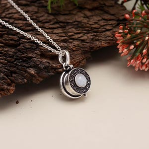 May include: A silver pendant necklace with a circular, decorative pendant. The pendant has a central, pale stone encircled by a ring of dark stones. The necklace chain is fine silver. The background is a mix of wood and a light surface.