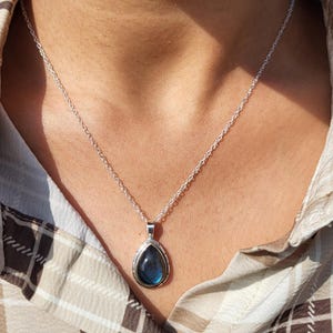 Puede incluir: Un collar de plata con un colgante en forma de lágrima. El colgante presenta una piedra preciosa azul con un borde plateado. El collar se lleva sobre una camisa a cuadros.