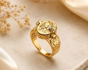 May include: A gold-toned ring featuring a hinged circular locket top with an embossed winged creature design. The band and the locket edges display a textured, weathered finish, with a small metallic accent visible on the hinge mechanism.