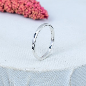 Op de afbeelding: Een glanzende, zilveren ring met een gladde, afgeronde band. De ring ligt op een wit oppervlak, met een wazig roze bloemelement op de achtergrond. De ring is een eenvoudige band, mogelijk een trouwring of stapelring.
