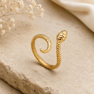 Puede incluir: Un anillo de serpiente dorado con una superficie texturizada. La cabeza de la serpiente está detallada con pequeñas hendiduras, y el cuerpo se curva en espiral. El anillo está sobre un fondo neutro.