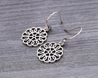 May include: A pair of silver-colored dangle earrings. Each earring features a circular, openwork design with a floral pattern. The earrings have a simple hook closure and are displayed on a gray wooden surface.