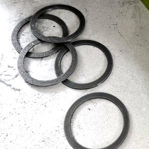 May include: A collection of dark gray, circular gaskets with a textured surface. The gaskets are arranged in a slightly overlapping pattern, with one gasket isolated at the bottom. The background is a light gray, speckled surface.