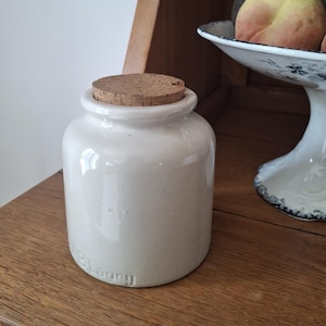 Peut inclure: Un petit pot en céramique blanc cassé avec un bouchon en liège. Le pot a un corps arrondi et un bord légèrement évasé. Le mot "Lagny" est estampé sur le côté. Le pot est posé sur une surface en bois.