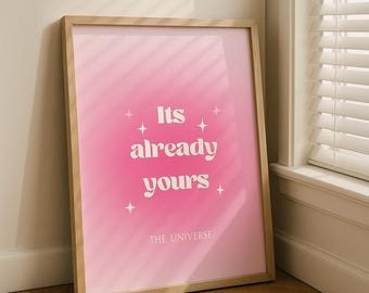 May include: A framed print with a pink gradient background and the words "Its already yours" in white, with the text "-THE UNIVERSE" below. The frame is made of light-coloured wood. The print is leaning against a wall.