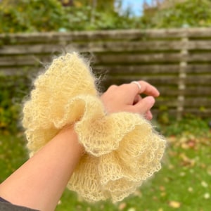 Könnte beinhalten: Ein großer, flauschiger, hellgelber Scrunchie aus weichem, strukturiertem Material. Der Scrunchie hat ein Rüschendesign und wird am Handgelenk getragen. Der Hintergrund ist eine verschwommene Ansicht einer grünen, äußeren Umgebung.