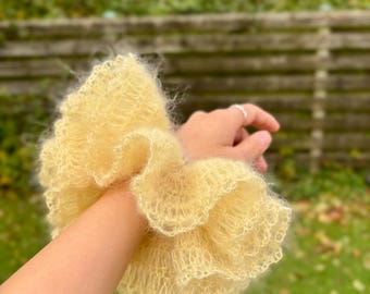 May include: A large, fluffy, pale yellow scrunchie made of a soft, textured material. The scrunchie has a ruffled design and is worn on the wrist. The background is a blurred view of a green, outdoor setting.