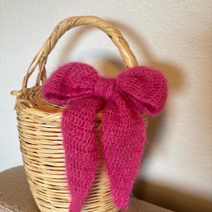 Peut inclure: Un petit panier en osier tressé avec une anse incurvée. Un grand nœud rose vif et duveteux est attaché à l'avant du panier. La texture du nœud contraste avec le motif tressé du panier.