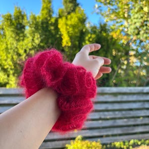 May include: A vibrant red scrunchie made of fluffy, textured material. The scrunchie is wrapped around a wrist, showcasing its voluminous, gathered design. The accessory adds a pop of color and texture.