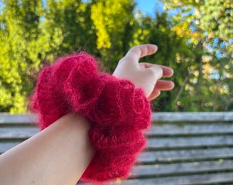 May include: A vibrant red scrunchie made of fluffy, textured material. The scrunchie is wrapped around a wrist, showcasing its voluminous, gathered design. The accessory adds a pop of color and texture.