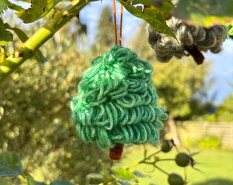 Op de afbeelding: Een close-up van een handgemaakte, groene gehaakte kerstboomversiering die aan een tak hangt. De boom heeft een bruine stam en wordt opgehangen met een bruin touwtje. Een andere, vergelijkbare versiering is zichtbaar op de achtergrond.
