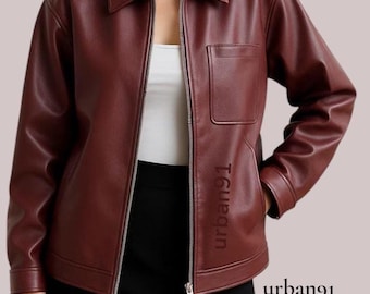 Puede incluir: Chaqueta burdeos con cuello y cremallera frontal. Un bolsillo en el pecho y el texto "urban91" impreso en la manga. La persona lleva una falda negra y una camiseta blanca. La chaqueta es de color rojo oscuro.