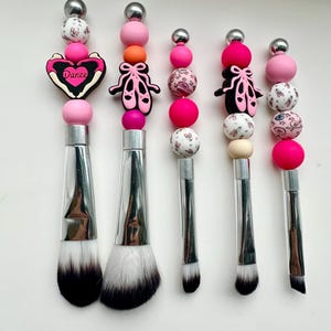 May include: Five makeup brushes with silver handles and white and black bristles. Each brush features a beaded design in pink, white, and silver. Some beads have ballet-themed charms and the word "Dance".