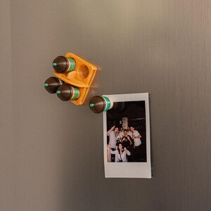 Op de afbeelding: Een houten kruidenrek met vier kruidencontainers en een enkele Polaroid-foto bevestigd aan een grijs oppervlak. De kruidencontainers zijn bruin met groene en witte banden. De Polaroid-foto toont een groep mensen.