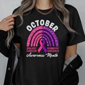 A person wearing a black t-shirt with a graphic design featuring a pink and purple rainbow, a pink ribbon symbol, and text related to breast cancer awareness.
