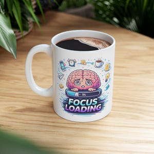 Puede incluir: Taza de café de cerámica blanca con asa, llena de café oscuro. La taza presenta un gráfico de cerebro de dibujos animados con el texto "FOCUS LOADING" y otras pequeñas ilustraciones. La taza está sobre una superficie de madera.
