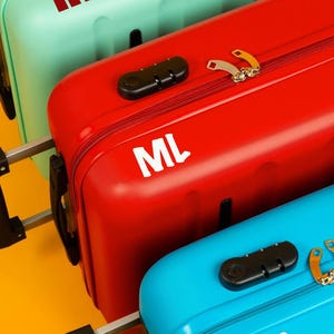 May include: Three colorful suitcases in mint green, red, and blue, each with a combination lock and a silver handle. The red suitcase has the initials "ML", while the other two have "HF".
