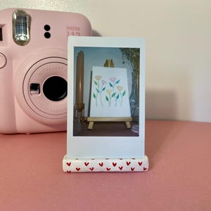 Könnte beinhalten: Eine hellrosa Instax 12 Sofortbildkamera neben einem weißen, rechteckigen Ständer, der mit kleinen roten Herzen verziert ist. Der Ständer hält ein vertikales Foto, das eine Miniatur-Holzstaffelei mit einer Zeichnung von Blumen zeigt.