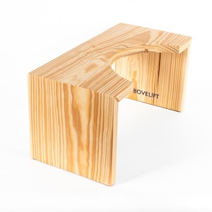 Puede incluir: Un taburete de madera con forma rectangular y un recorte curvo. El taburete está hecho de madera clara con vetas visibles. La palabra "BOVELIFT" está impresa en el lateral.