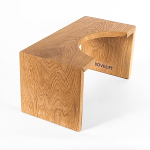 May include: A wooden footstool with a natural wood grain finish. The stool has a rectangular top with a curved cutout and two supporting legs. The word "BOVELIFT" is printed on the side. The stool is made of solid wood.