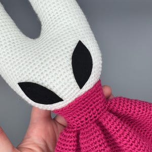 Könnte beinhalten: Handgefertigtes Häkeltier mit weißem Kopf, schwarzen Augendetails und einem pinkfarbenen Körper. Die Textur des Spielzeugs wird durch sichtbare Maschen definiert. Das Design ist von einer Videospiel-Figur inspiriert.