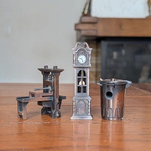 Op de afbeelding: Drie miniatuur metalen beeldjes waaronder een donkerbronskleurige drukpers, een hoge staande klok met een witte wijzerplaat en een kleine cilindrische prullenbak met deksel, allemaal rechtopstaand op een vlak houten oppervlak.