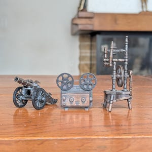 Puede incluir: Tres objetos de colección en miniatura de metal con acabado en bronce oscuro. El conjunto incluye un pequeño cañón de artillería con dos ruedas, un proyector de cine de estilo vintage con dos bobinas circulares y una rueca alta y detallada.