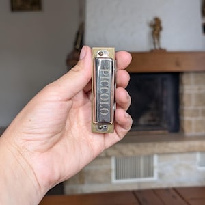 May include: A silver and gold harmonica with the word "Piccolo" printed vertically. The instrument is held in a hand, with a blurred background of a fireplace and wooden beams.