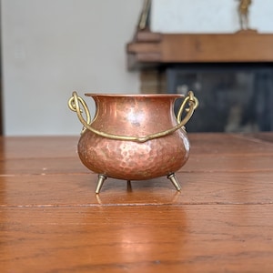 May include: A small, round, hammered copper pot featuring a brass swing handle and three short brass legs. The pot has a wide, open top and a textured, metallic surface, showcasing a warm, reddish-brown finish with contrasting gold-toned metal accents.