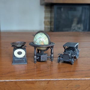 Puede incluir: Una colección de tres objetos metálicos en miniatura que incluye una báscula de cocina gris oscuro con esfera blanca, un pequeño globo terráqueo sobre un soporte de metal oscuro y un modelo de automóvil antiguo gris oscuro con cuatro ruedas.