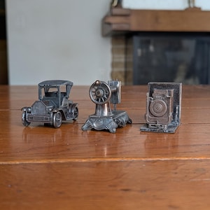 Könnte beinhalten: Drei metallene Miniatur-Sammlerstücke in einer Reihe. Das erste ist ein Automobil im Vintage-Stil, das zweite eine klassische Nähmaschine mit einem markanten Rad und das dritte eine antike Balgenkamera mit einem quadratischen Rahmen.