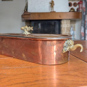 May include: A rectangular copper container with a lid and decorative brass handles and accents. The container is sitting on a wooden table. The lid has a fish-shaped handle. The handles on the sides are dragon-shaped.