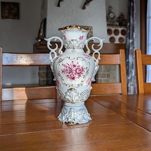 May include: An ornate white vase with gold trim and pink floral patterns, standing on a wooden table. The vase features decorative handles and a detailed base, showcasing its elegant design. The vase is a decorative item.