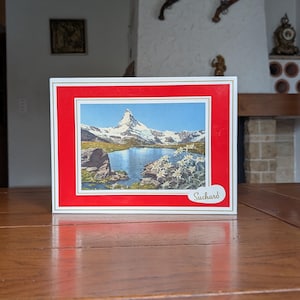 May include: A rectangular Suchard box with a red border features a scenic image of a snow-capped mountain reflected in a lake. The box is set on a wooden table, with a blurred background of a room.