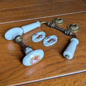 Puede incluir: Juego de herrajes de puerta antiguos con manijas de porcelana blanca con diseños florales, detalles de latón y rosetas decorativas. El juego incluye dos manijas y dos rosetas, ideal para proyectos de restauración.