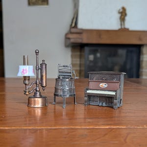 Puede incluir: Tres sacapuntas de metal en miniatura que incluyen una lámpara de aceite color cobre con pantalla blanca floral, un escurridor de ropa vintage plateado y un piano vertical marrón con teclado blanco y un pequeño logotipo circular.