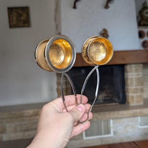 May include: Two tea infusers with silver-toned exterior handles and frames, featuring circular perforated bowls with a gold-toned interior finish. Each infuser has a hinged mechanism that allows the two halves of the bowl to open and close securely.