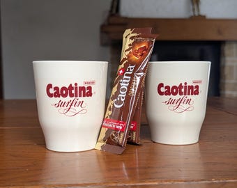 May include: Two white ceramic mugs with the Caotina Surfin logo in red script sit on a wooden table, alongside two packets of Caotina chocolate drink mix. The packets are brown with gold accents and the Caotina logo.