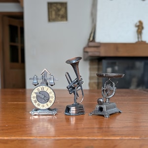 Könnte beinhalten: Drei Miniatur-Bleistiftspitzer aus Metall in Form einer Uhr mit zwei Glocken, einer messingfarbenen Trompete und einer Waage im Vintage-Stil. Jeder Gegenstand weist feine Details und ein metallisches Finish auf und steht auf einer ebenen Fläche.